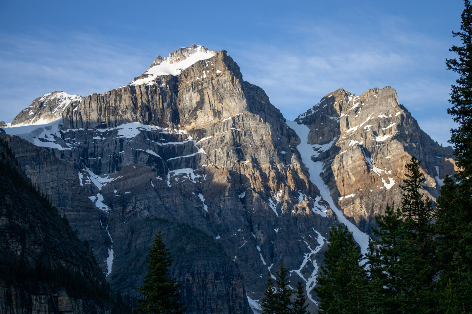 A telephoto view of a layered limestone peak with snow-filled couloirs, sidelit by early morning sun against a deep blue sky.