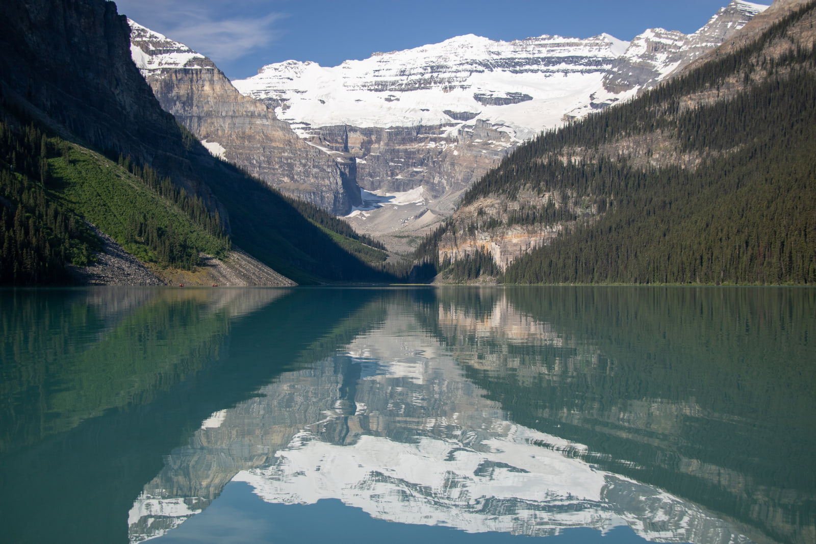 Mount Victoria and its glacier reflected in the mirror-calm turquoise water of Lake Louise, framed by forested ridges under a clear sky.