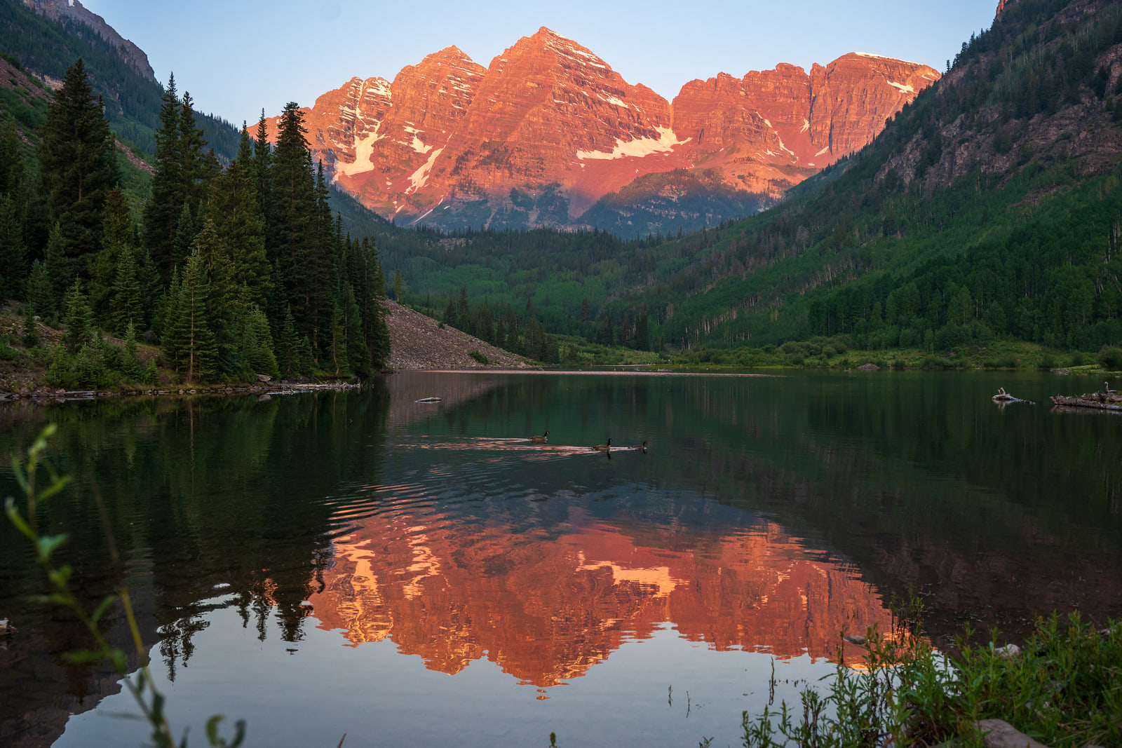 Twin mountain peaks glowing red in sunrise light are reflected in the still surface of an alpine lake framed by spruce trees.