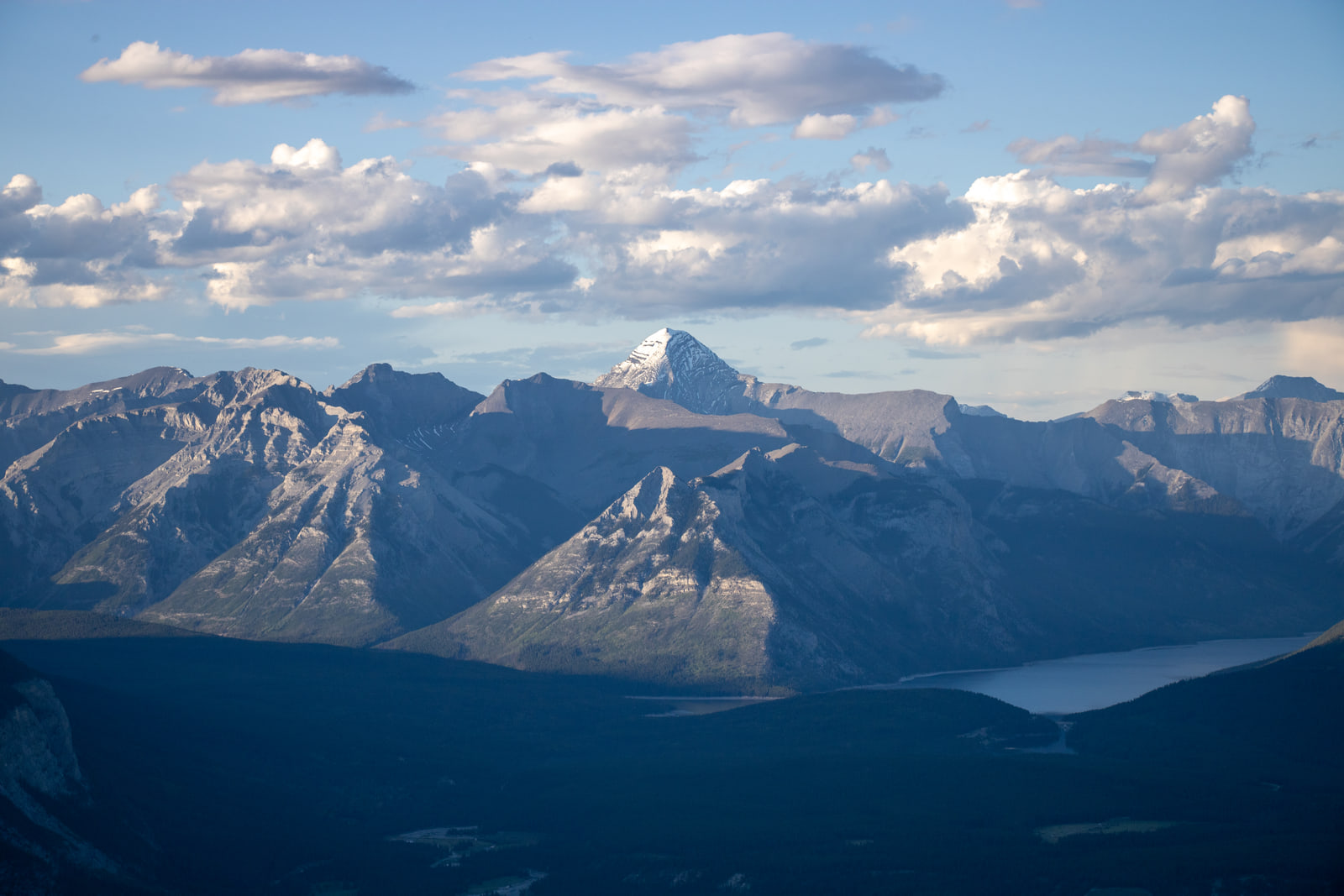 A snow-capped mountain peak rises behind layered grey ridges above a valley in deep shadow, with a sliver of lake visible at right under a sky of sunlit clouds.