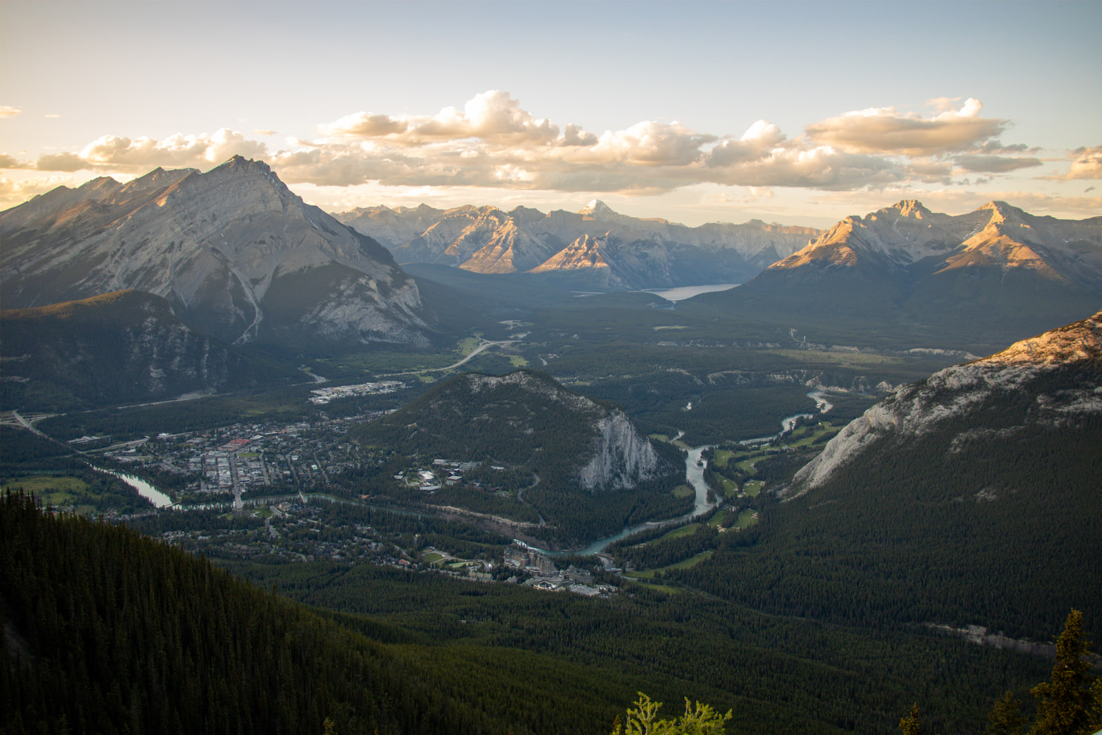 The town of Banff sits on a broad valley floor seen from high above, with the Bow River curving past a rocky outcrop and golden evening light on the surrounding mountain ranges.