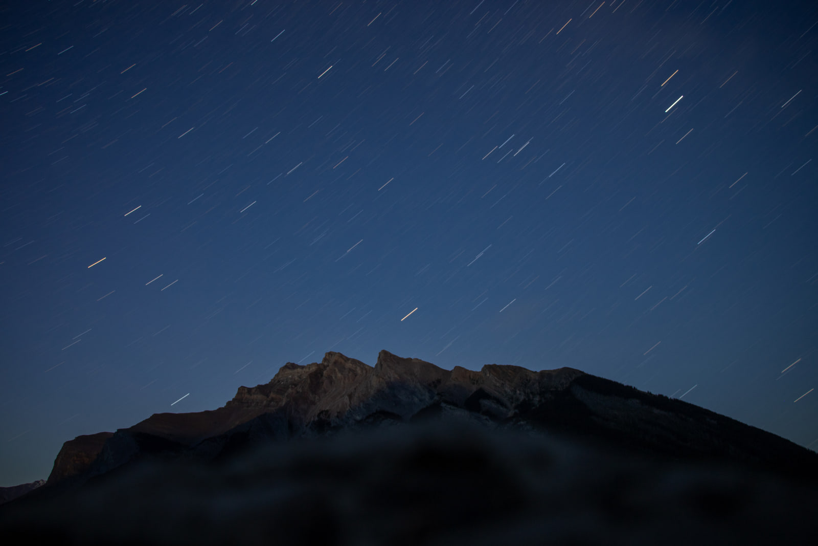 Short star trails streak diagonally across a deep blue night sky above the dark, faintly moonlit summit ridge of a mountain.