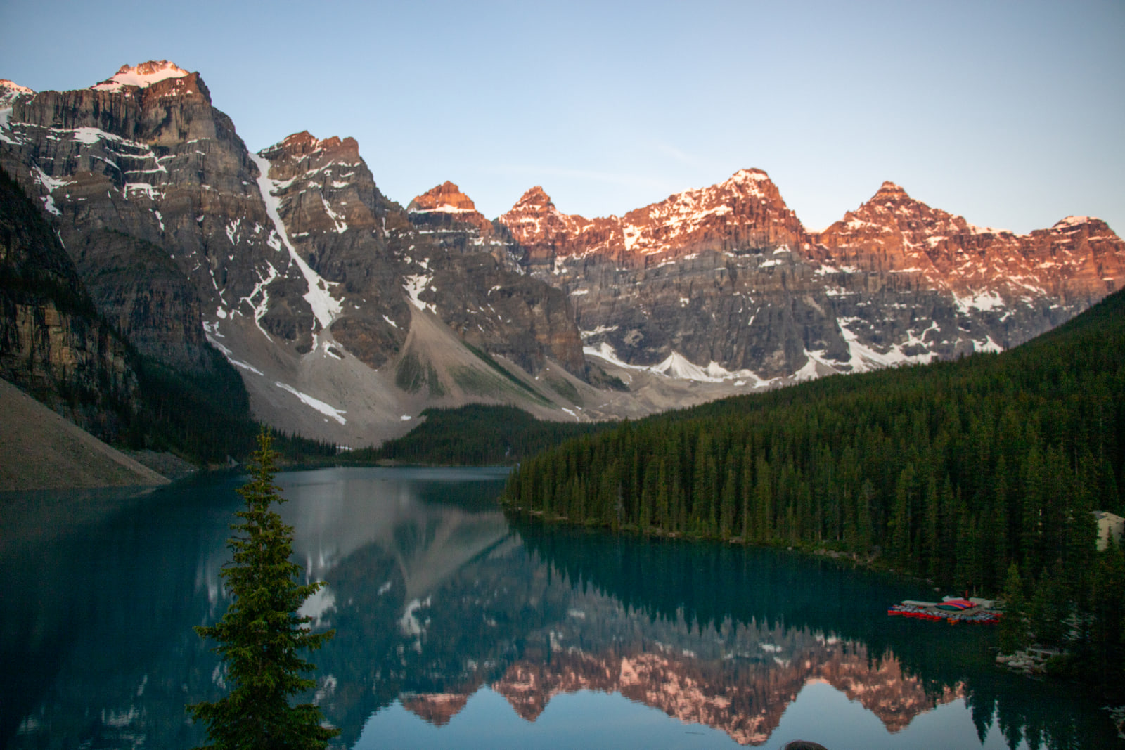 Pink sunrise light on the jagged Ten Peaks above Moraine Lake, the ridge reflected in still turquoise water with red canoes moored on the right shore.