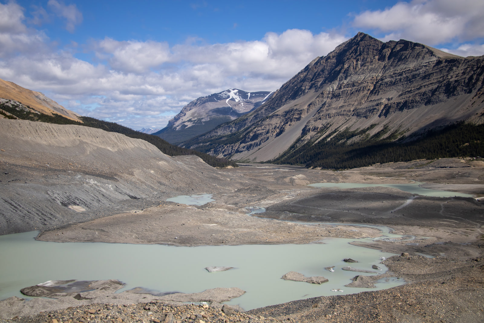 Turquoise meltwater pools with gravel islands fill a glacial valley below a dark ridge and a snow-capped peak.