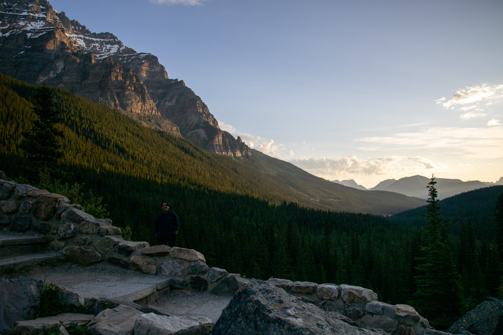 A lone figure seated on a stone trail wall above a forested valley, looking toward hazy sunlit ridges and low cloud in the distance.
